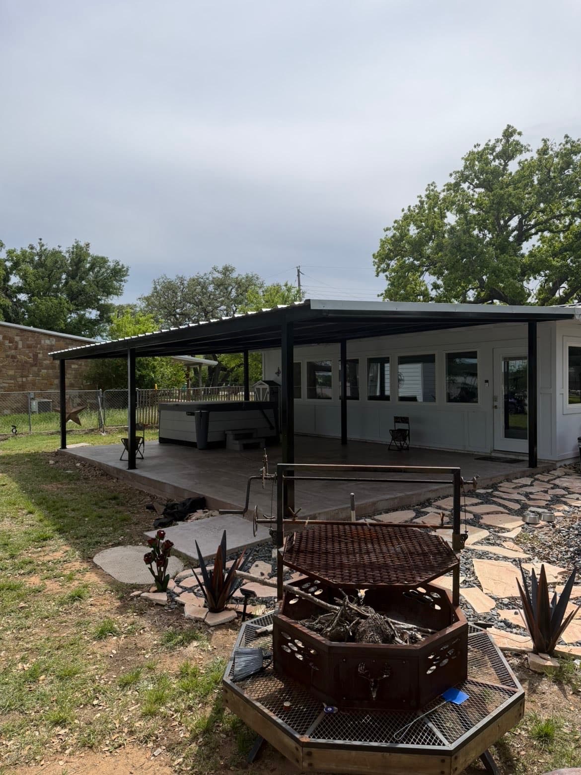 Finished black steel patio cover over a stained concrete slab behind a white house, with a steel fire pit in front