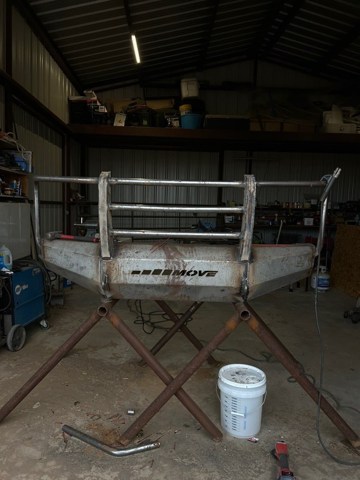 A custom steel grill guard and bumper on pipe stands in the shop, with a welder and tools along the wall