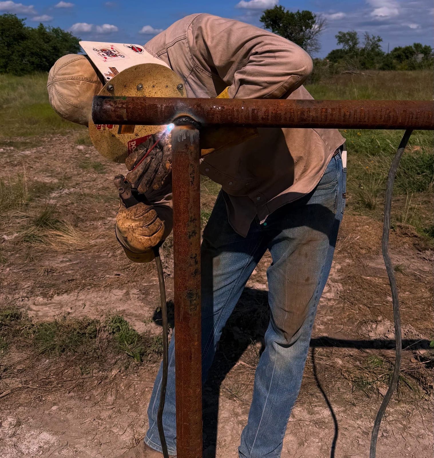 Welder joining a steel pipe rail to a fence post in an open pasture